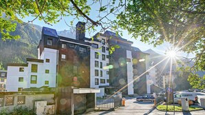 Un immeuble moderne à plusieurs étages, doté d’un parking à l’avant, entouré de verdure et de montagnes.