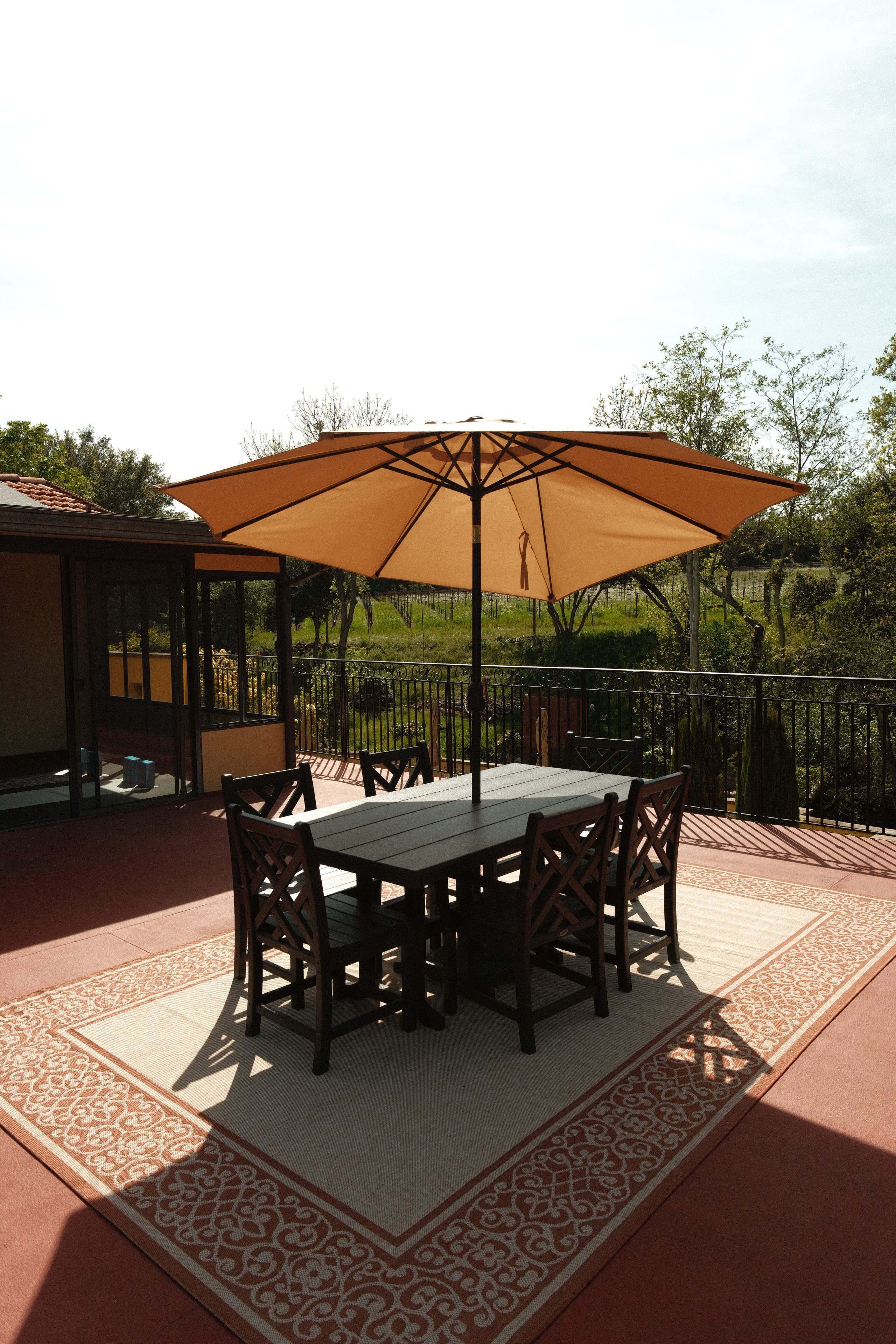 Une terrasse extérieure avec une table et des chaises sous un grand parasol, entourée d’un tapis à motifs et d’une clôture en métal.