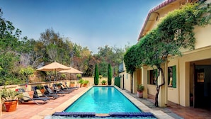 A rectangular pool with a tiled edge, surrounded by lounge chairs and umbrellas, with a building and trees in the background.