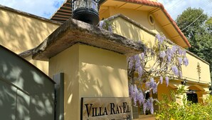 En villa kaldet "Villa Rayel" med en stenelegant indgang, et skilt der angiver stiftelsesår 1974, og lilla wisteria blomster.