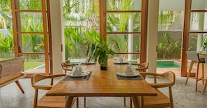 A wooden dining table with chairs, set for four, in a room with large windows overlooking a garden with various plants and a pool.