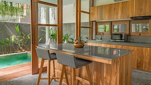 A modern kitchen with wooden cabinets, a black countertop, and a small island with bar stools. - Villa Avyathi at Anandathu Villas - Tropical 2BR Contemporary Villa in Canggu (Bali)