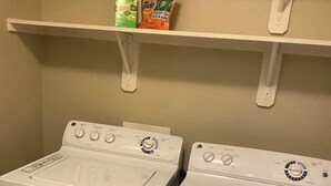Un espace buanderie équipé de deux machines à laver à chargement frontal et d’une étagère au-dessus, où sont rangées les bouteilles de lessive.