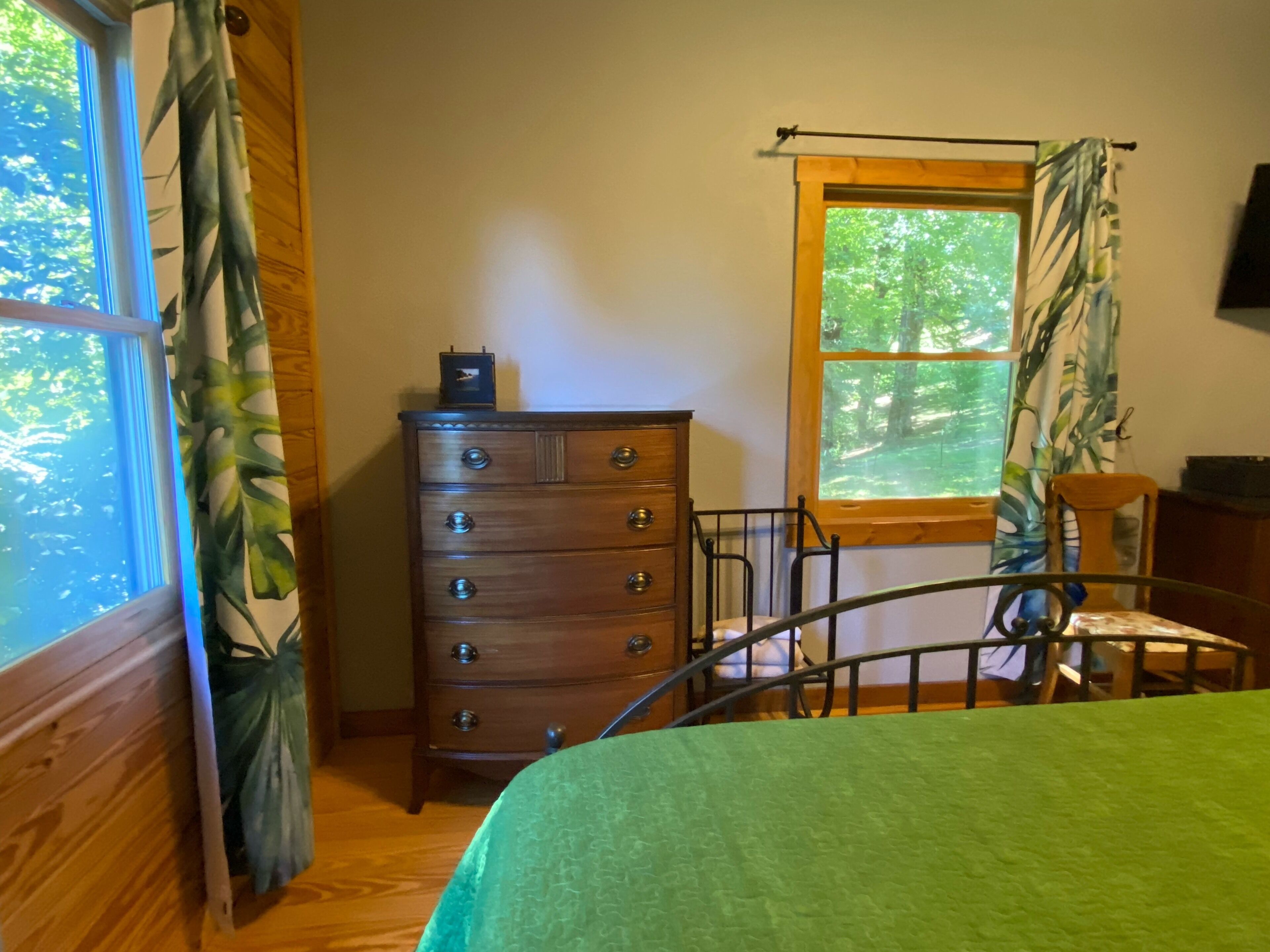 Een kamer met een houten dressoir, een metalen bed en een stoel, met uitzicht op bomen.