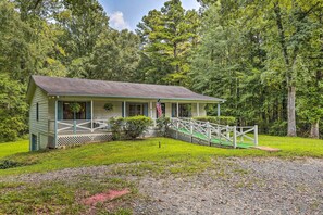 A small, single-story cabin with a porch, surrounded by lush greenery and a flag. - Quiet Home w/ Fire Pit - 1 Mi to Downtown Acworth! (Acworth)