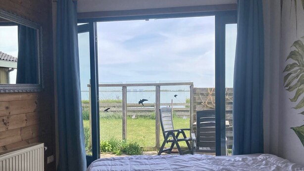 A bedroom with a large bed, a view of the outdoors, and a window with curtains. - 6 pers Holiday home Aurora with directly on the Lauwersmeer (Anjum)
