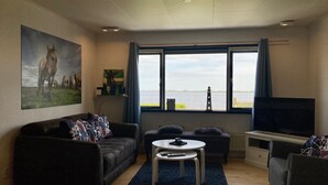 A living room with a sofa, a coffee table, a TV, and a large window with a view of the water. - 6 pers Holiday home Aurora with directly on the Lauwersmeer (Anjum)