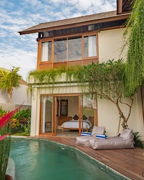 Una casa de dues plantes amb una porta de vidre que dona a un dormitori amb llit i sofà. Hi ha una piscina en primer pla.