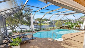 Et overdækket udendørs swimmingpool med liggestole og plantekasser, omgivet af et metalhegn med udsigt over en sø.