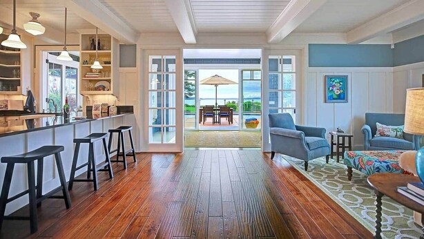 A spacious room with a wooden floor, a kitchen area with bar stools, a seating area with chairs and a table, and a view through large doors to an outdoor patio.