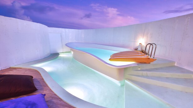 Una piscina interior moderna con diseño curvo, una pequeña zona de estar con cojín y una escalera que conduce a un nivel superior.