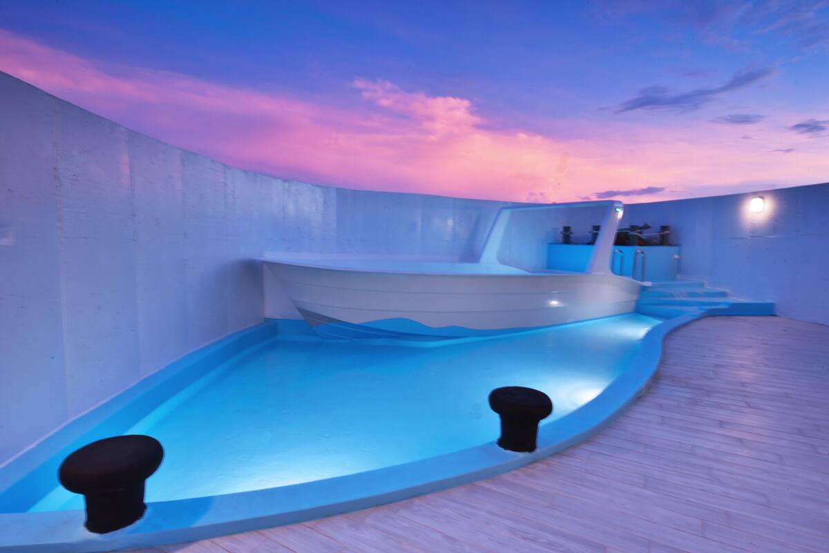 Piscina interna moderna con un design unico a forma di barca, circondata da un pontile in legno e un cielo rosato.