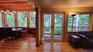 A wooden cabin interior with a dining area, a sofa, and large windows offering a view of the forest.