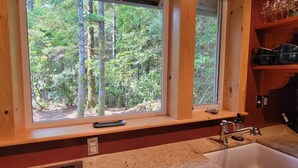 A kitchen with a large window overlooking a forest, a NINJA coffee maker, and wooden shelves with dishes.