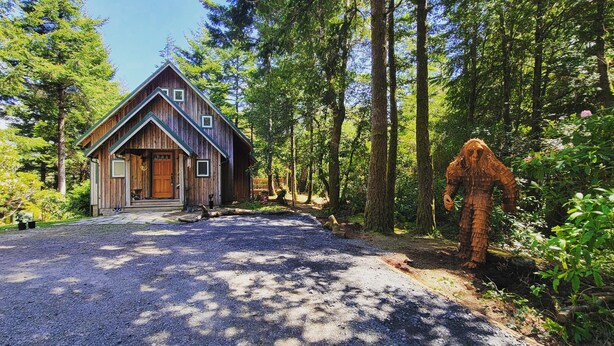 A rustic cabin with a steeply pitched roof, a front porch, and a gravel driveway.