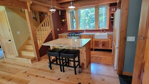 A wooden interior with a kitchen island, two stools, a stove, and a window overlooking greenery.