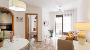 Un salon moderne comprenant un canapé, une table à manger avec des chaises, un ventilateur de plafond et un coin cuisine visible à travers une arche.