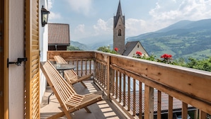 A wooden deck with a table and chairs, overlooking a church and green valley. - Holiday Apartment "Plose Winklerhof" with Mountain View, Balcony & Wi-Fi (Villanders)
