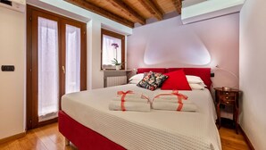 Une chambre avec un plafond en bois, un lit avec une literie blanche et des touches de rouge, une table de chevet et une fenêtre agrémentée d’une plante.