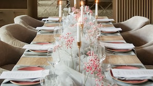 Una mesa de comedor dispuesta con manteles blancos, flores rosadas y velas.
