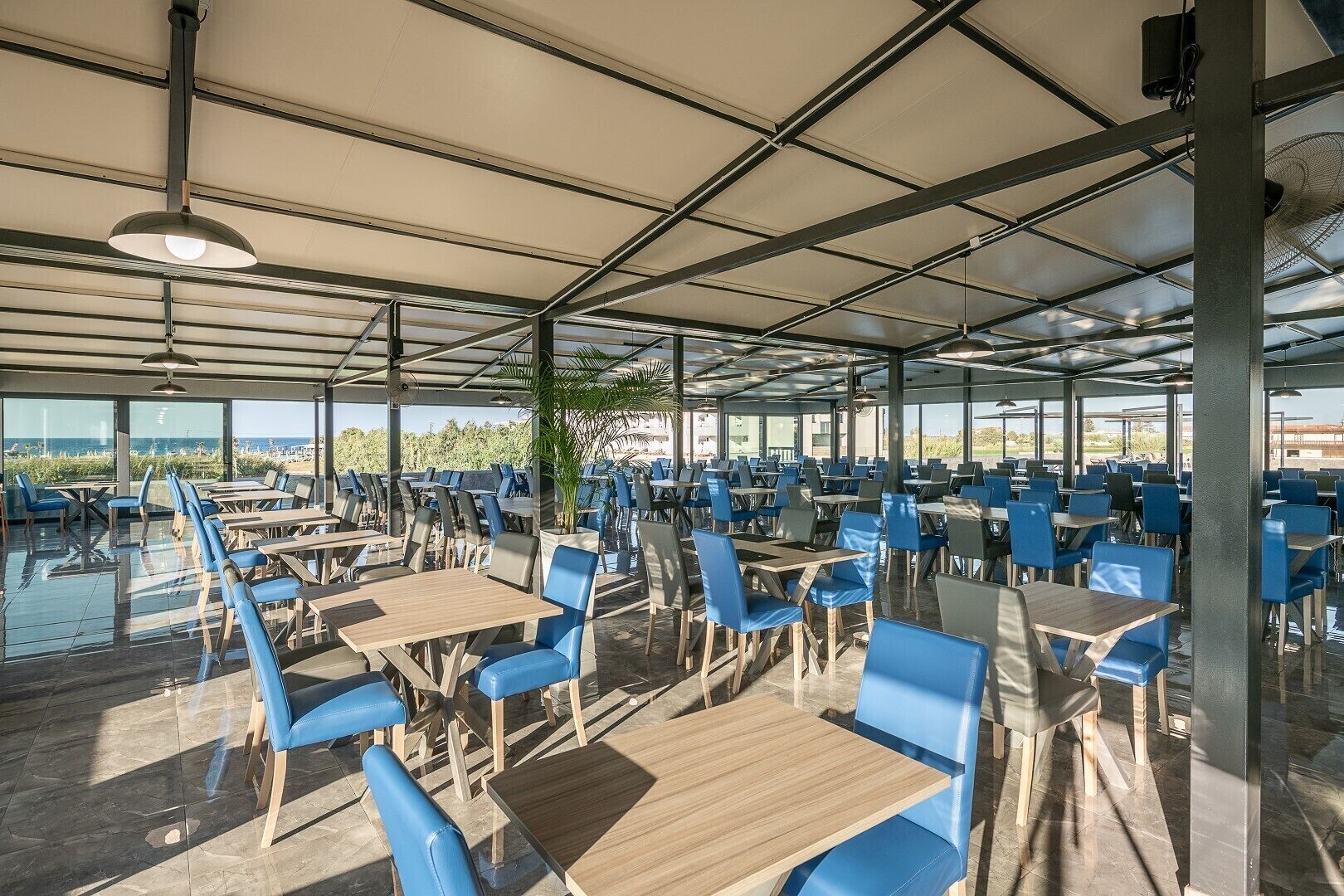 A spacious outdoor dining area with numerous tables and blue chairs, large windows offering a view of the beach, and a covered ceiling structure.