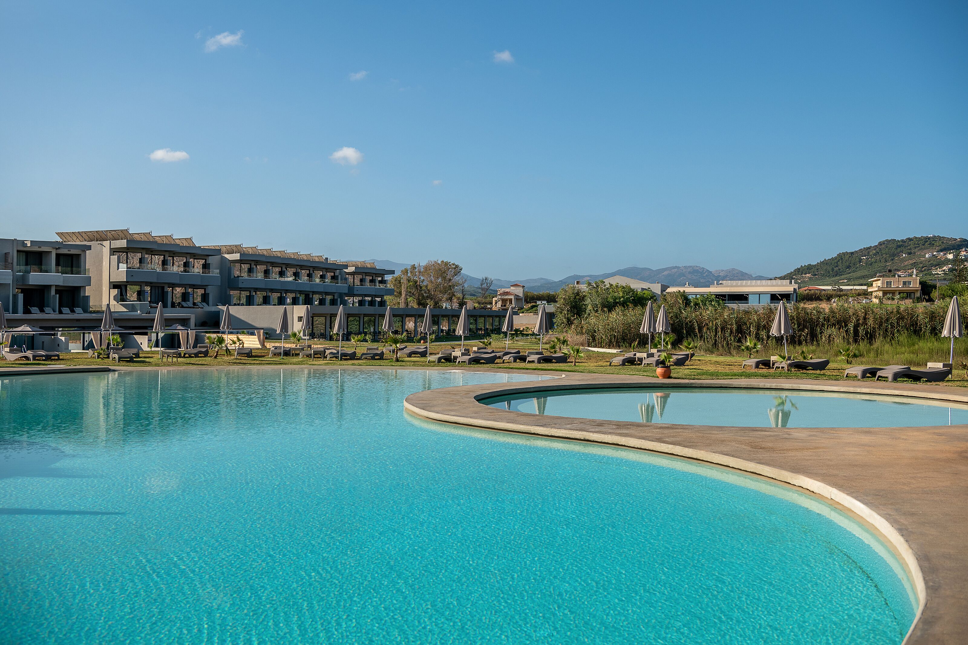 A swimming pool with lounge chairs and umbrellas, surrounded by buildings and a hilly landscape.