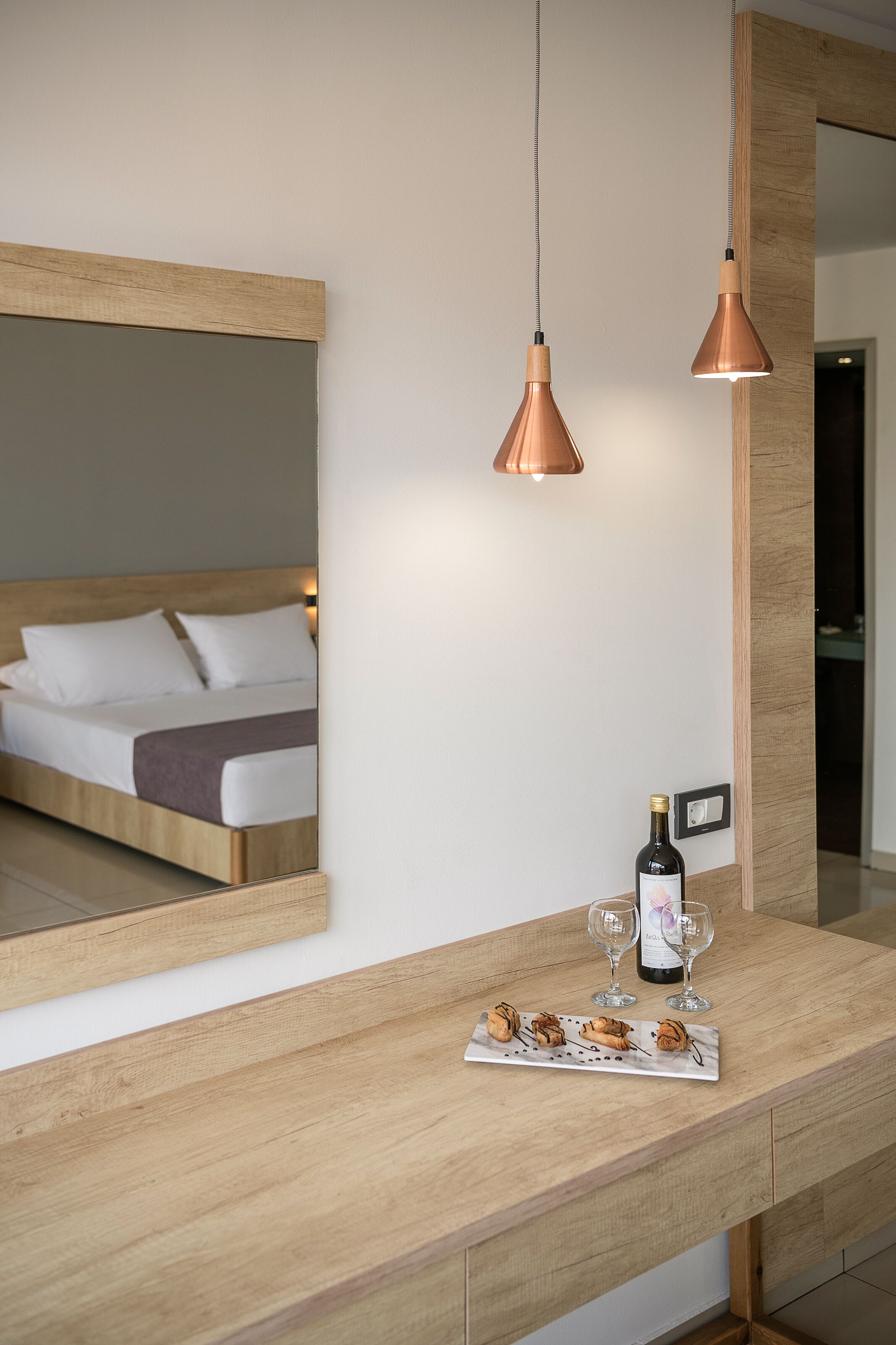 A modern hotel room with a wooden headboard bed, a large mirror, and a wooden console table with a bottle of wine and glasses.