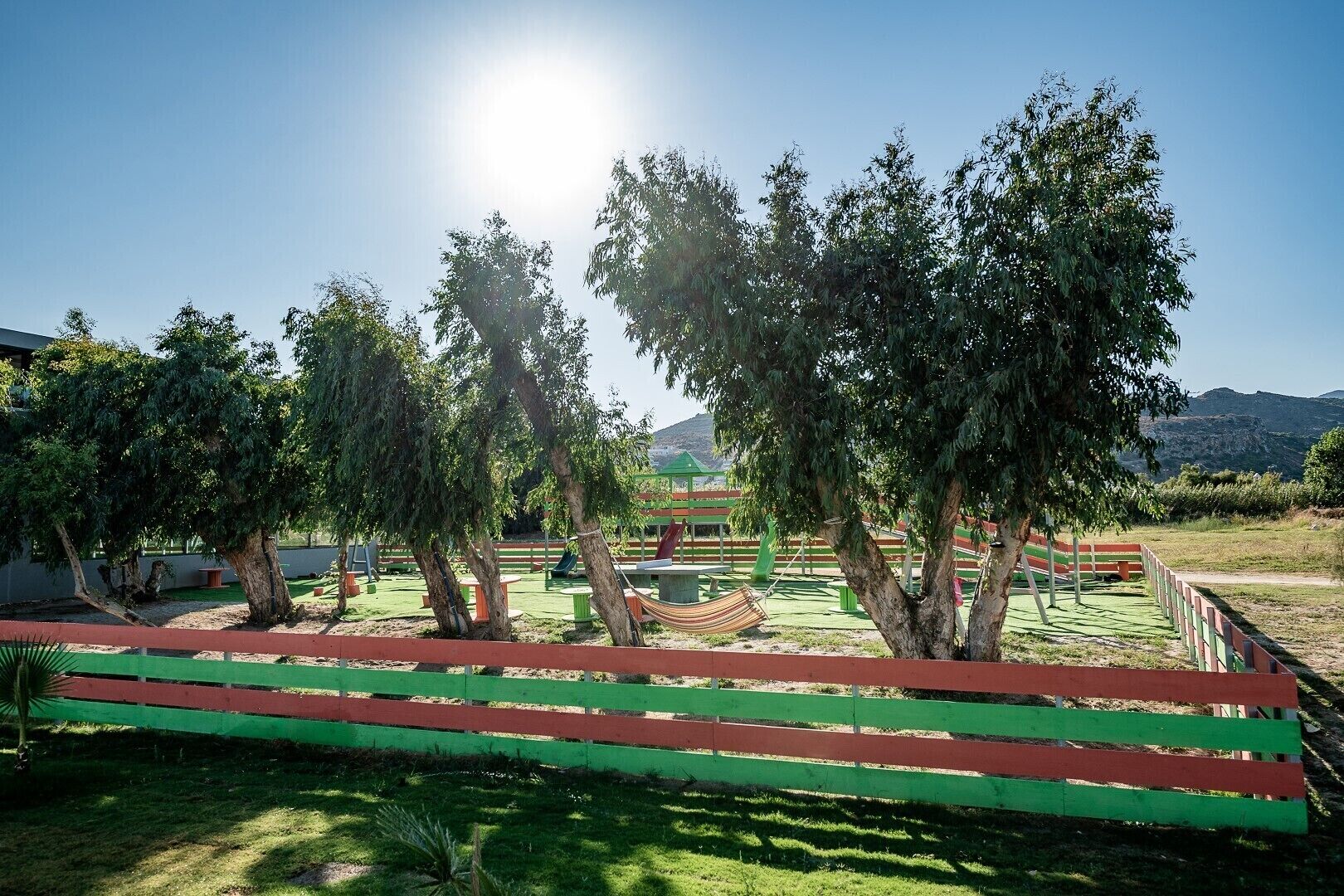 Un parque infantil con estructuras de juego verdes y rojas, rodeado de árboles y un cielo despejado.
