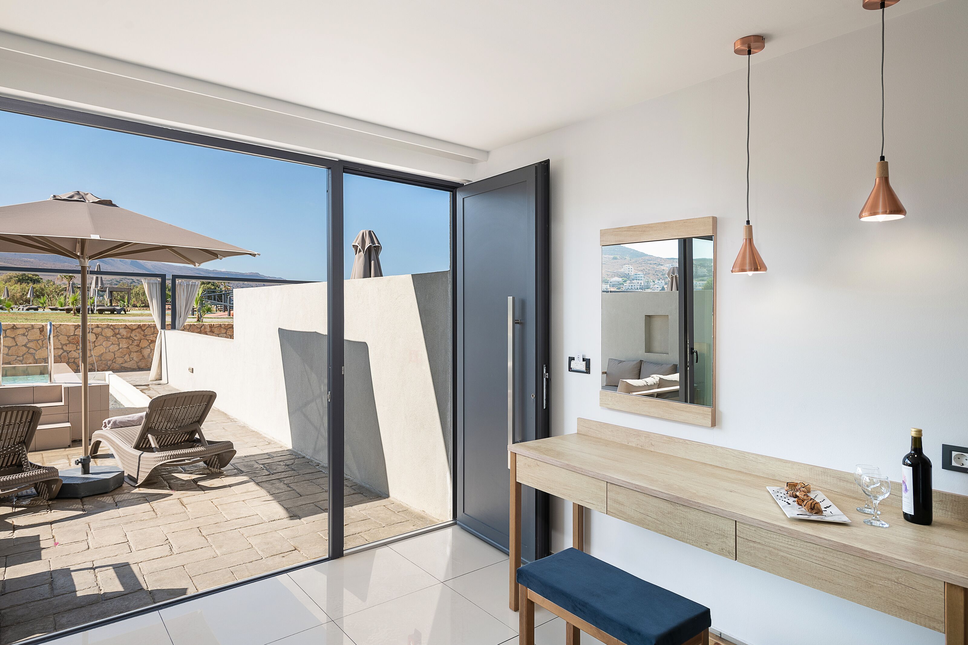 A modern room with a sliding glass door leading to an outdoor patio, a wooden bench, a table with a bottle and glasses, and a mirror on the wall.