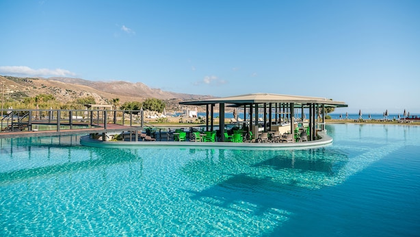 A floating bar with green chairs and umbrellas, surrounded by a pool with clear blue water, and a view of distant mountains.