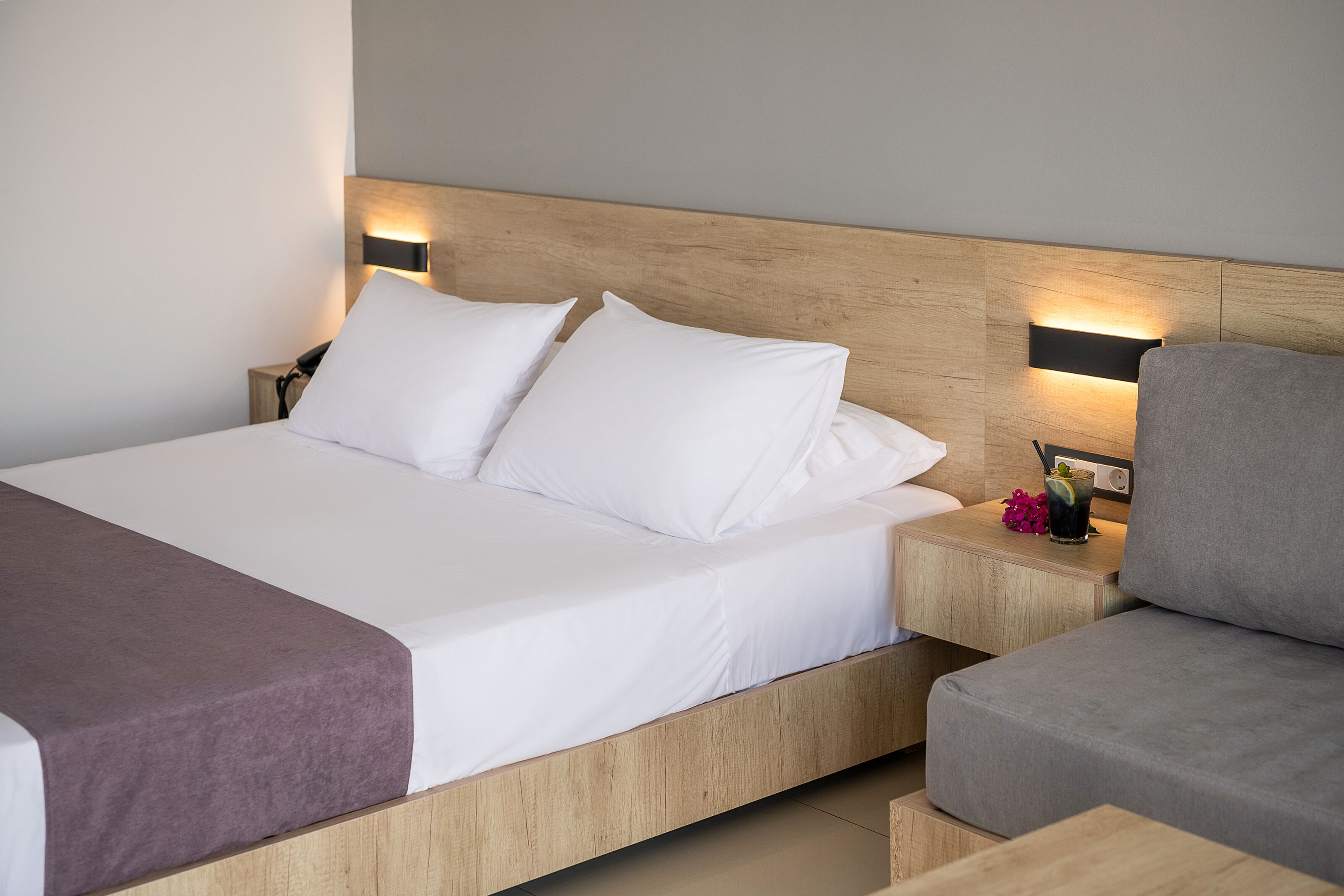 Una habitación de hotel moderna con cabecera de madera, una cama grande con ropa blanca, un sofá gris y mesitas de noche con lámparas.