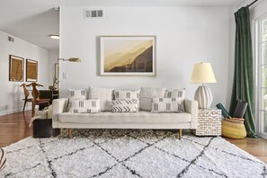 Una sala de estar con un sofá, cojines estampados, un cuadro de paisaje enmarco, una lámpara y una alfombra.
