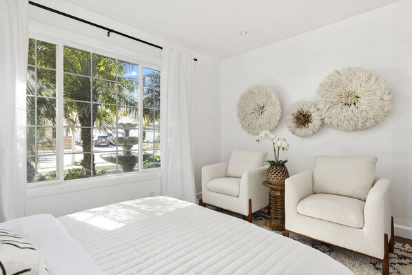 Un dormitorio con un ventanal grande, dos sillones, una cama y decoración en la pared.