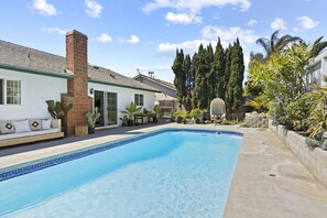 Una piscina con borde de azulejos, rodeada por un patio con mobiliario exterior y plantas.