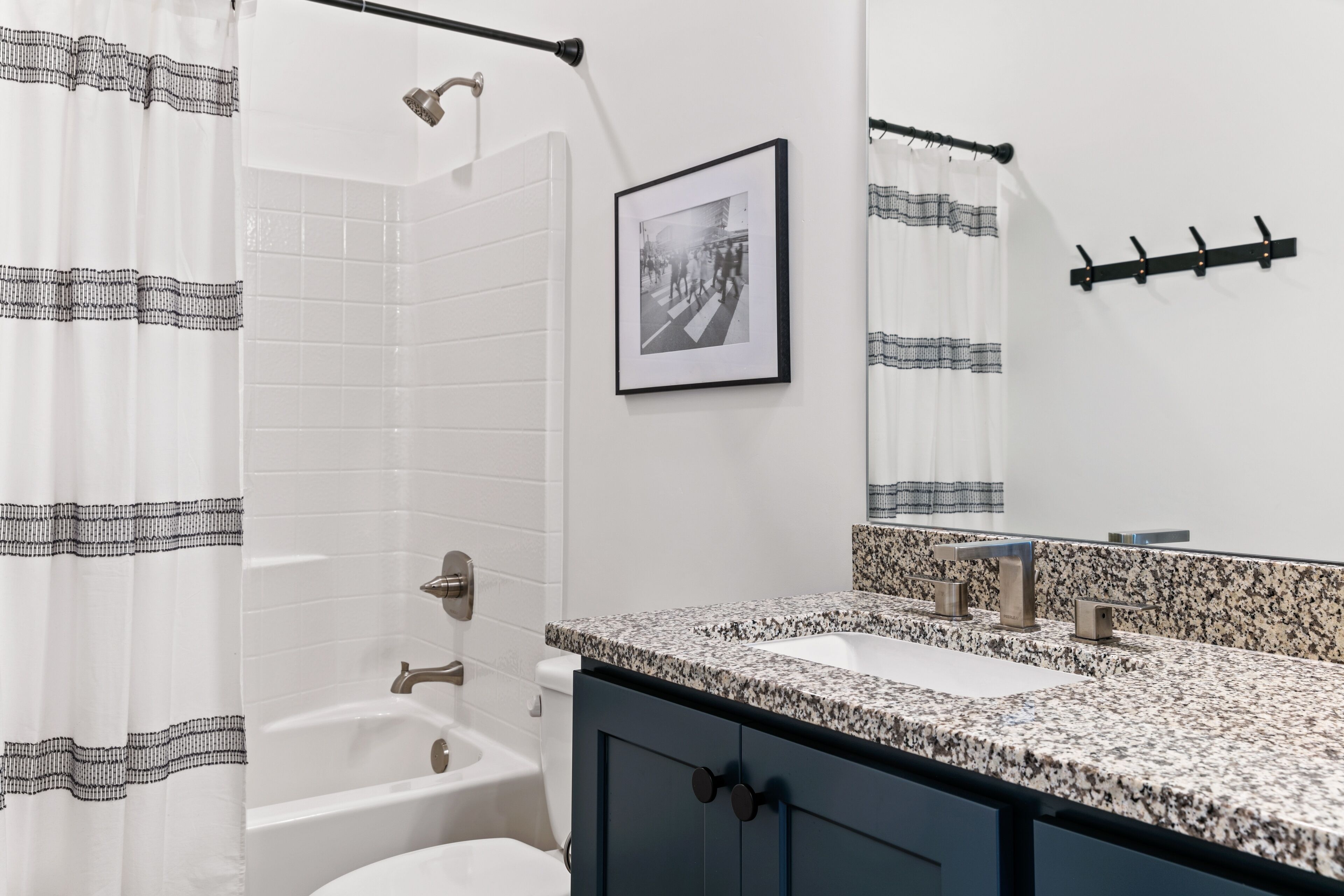 A bathroom with a white tiled wall, a shower with a black and white patterned curtain, a framed photograph on the wall, a granite countertop, and a sink with a faucet.