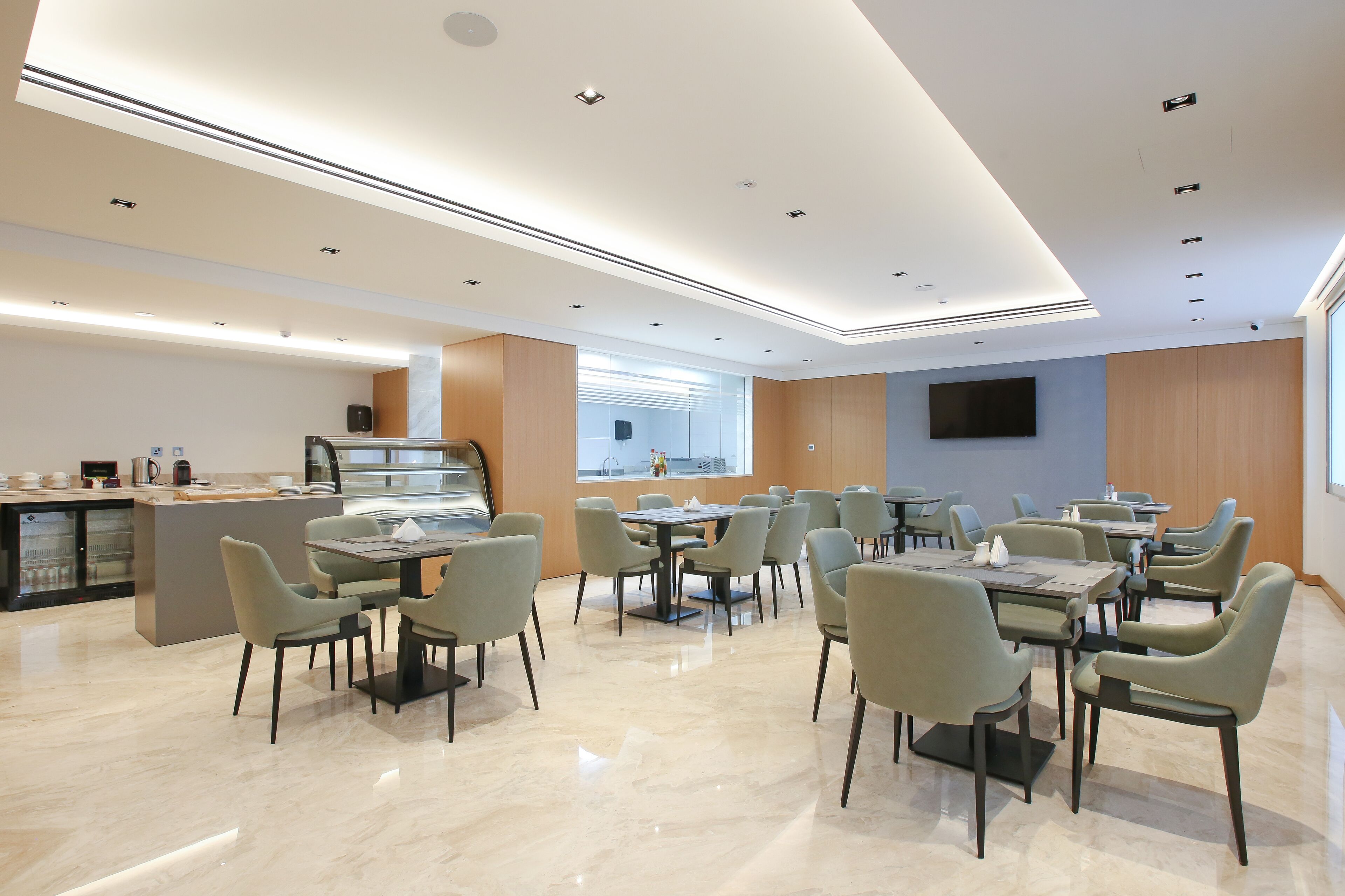 Un espace repas moderne avec plusieurs tables et chaises, un coin cuisine doté d’un meuble vitrine et une grande fenêtre qui laisse entrer la lumière naturelle.