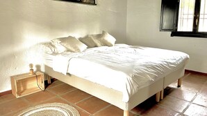Cama bem arrumada com lençóis brancos, abajur e tapete tecido sobre piso de azulejos.