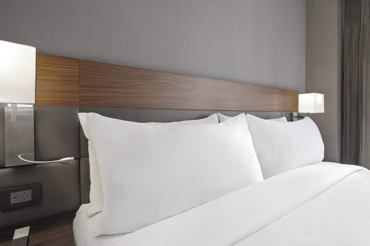 A hotel room with a wooden headboard, white bedding, a bedside table with a lamp, and a power outlet.