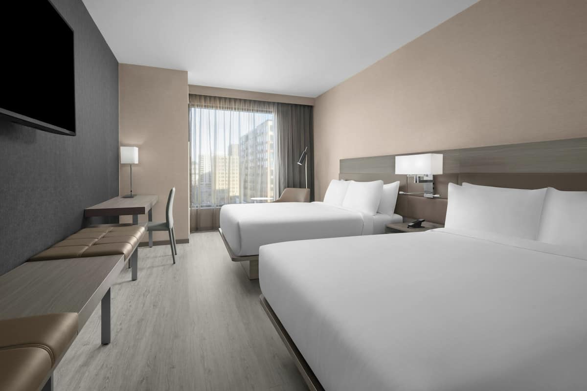 Habitación de hotel con dos camas, un escritorio, una silla, un televisor y un ventanal con cortinas.