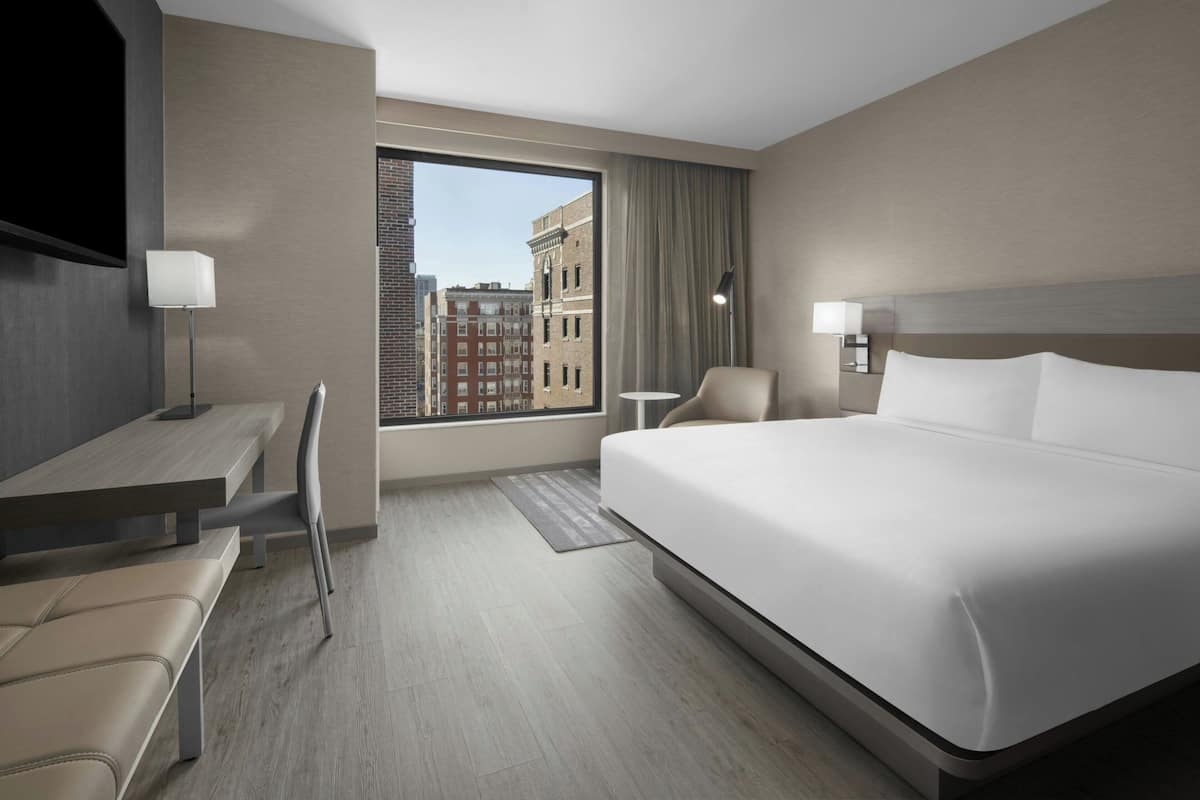 Habitación de hotel con una cama grande, un escritorio con silla, una mesita y vistas a la ciudad a través de un ventanal amplio.