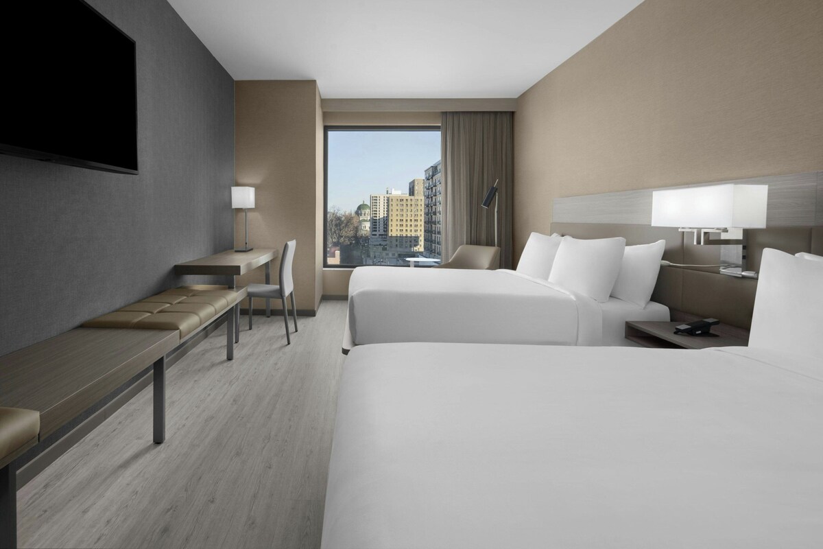 A hotel room with two beds, a desk, a chair, a TV, and a large window offering a city view.