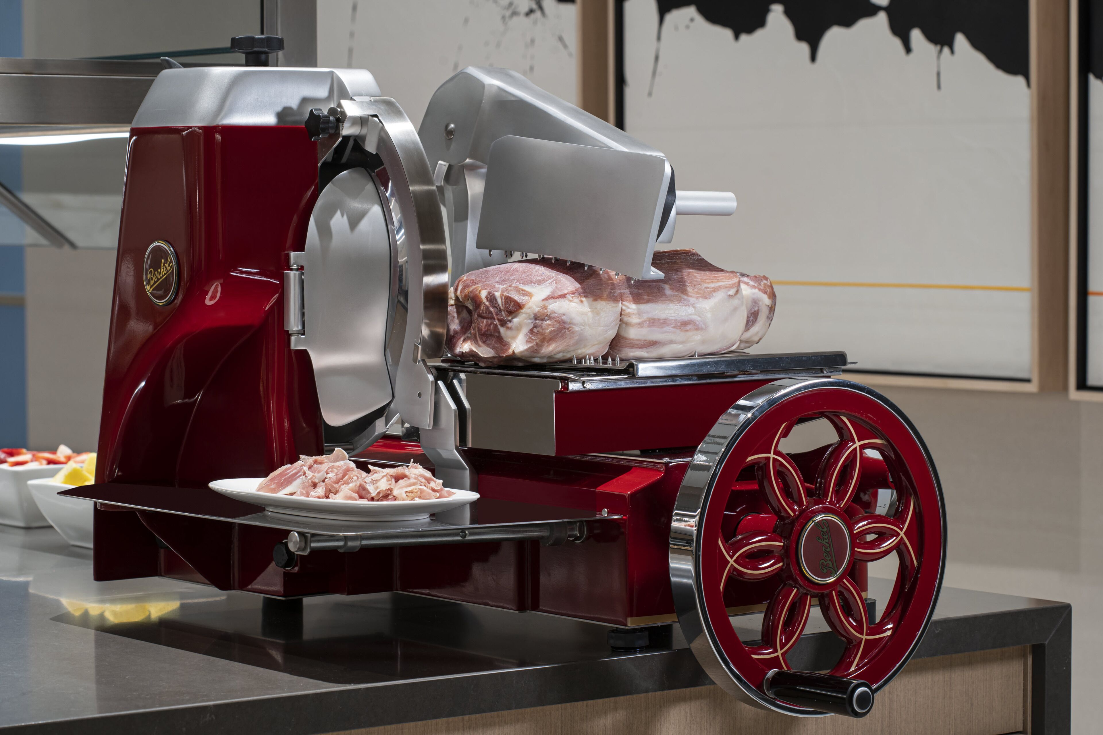 A meat slicer with a red and silver design, a plate of sliced meat, and a bowl of raw meat on a counter.