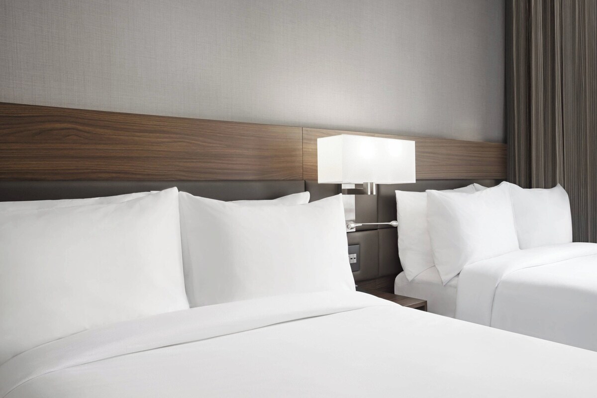 A hotel room with a wooden headboard, two beds with white linens, a bedside table with a lamp, and a grey curtain.