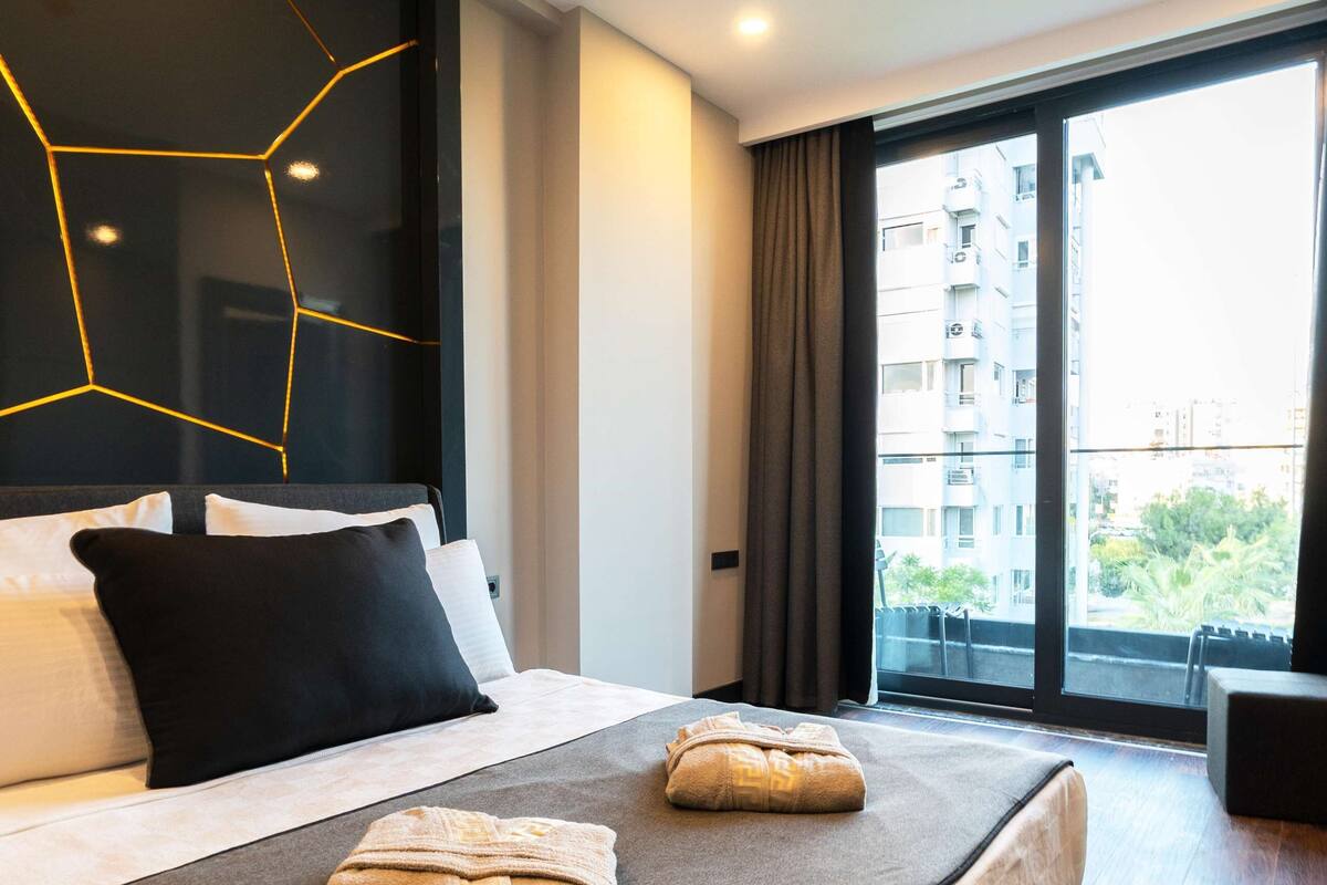 A modern hotel room with a large bed, a view of the city through large windows, and a geometric light fixture on the wall.