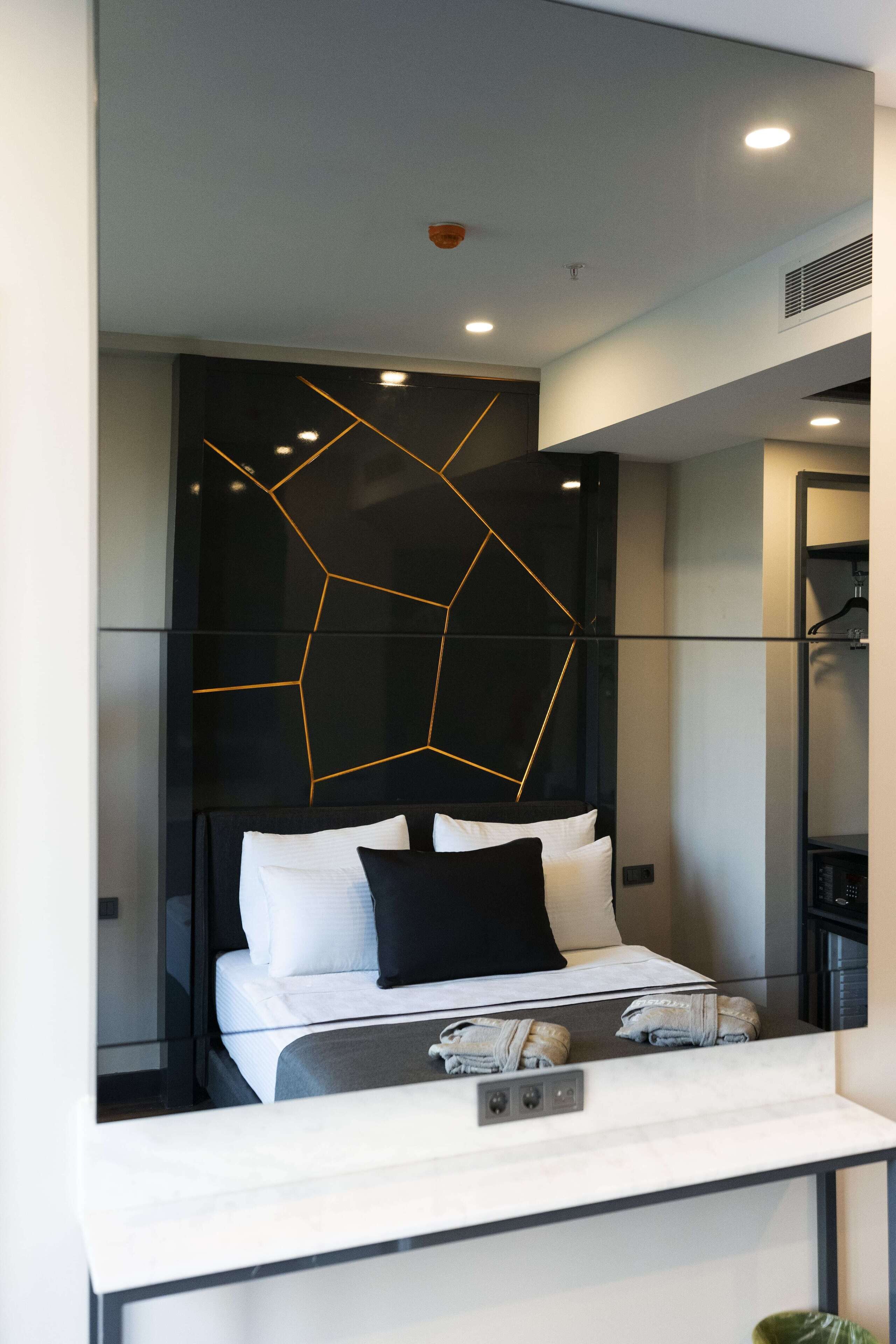 Une chambre moderne avec un motif géométrique sur le mur, un lit avec une literie blanche et noire, et une petite table de chevet avec une lampe.