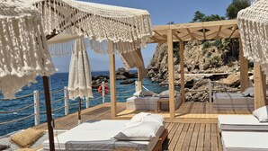 Une terrasse en bois, agrémentée de lits blancs et de parasols en macramé, qui offre une vue sur un paysage rocheux et la mer.