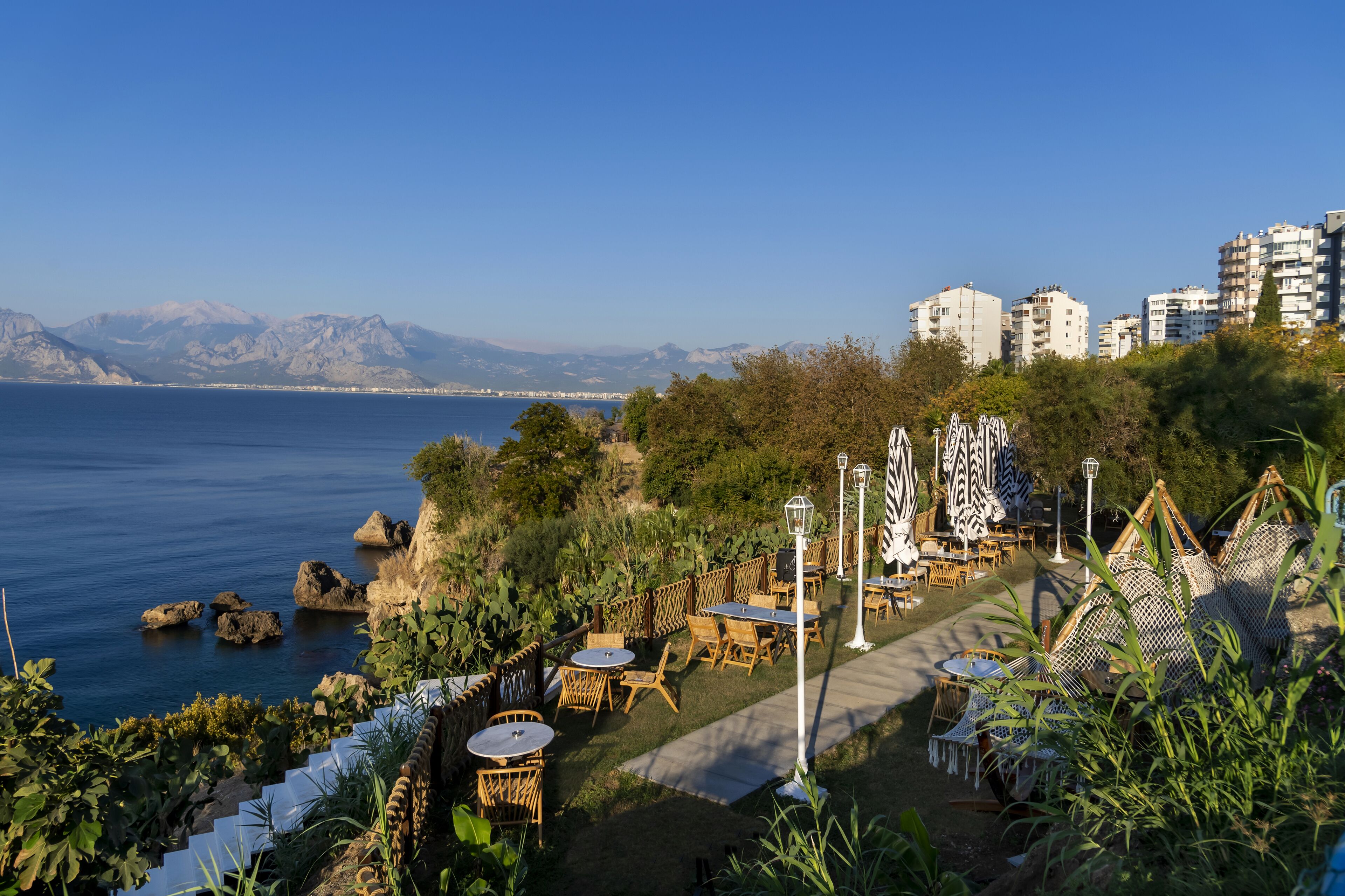 A coastal outdoor seating area with tables and chairs, overlooking the sea and mountains.