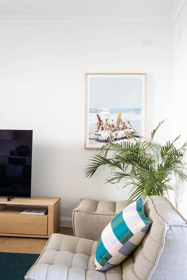 A living room with a television on a wooden stand, a beige sofa with a striped pillow, and a framed picture of a beach scene on the wall.