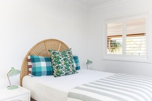 A bedroom with a bed, a wicker headboard, a window with blinds, and a bedside lamp.