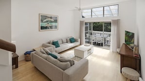 A modern living room with a sofa, coffee table, and a TV. There is a balcony with a view of the outdoors.